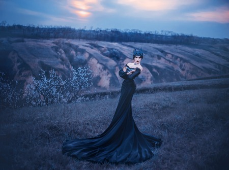 grim lady with pale white skin and dark hair beside flowering tree, witch turns with black crow with long luxurious dress and open shoulders, gothic image and makeup, cold metal crown and jewelsの写真素材