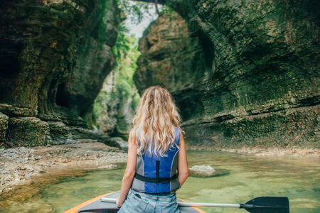 girl with blond hair in boat on lake in Georgia, nature Martvili Canyon, active leisure for tourists, gorgeous bright nature, tourist rafting entertainment on water, photo without face from backの写真素材
