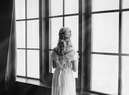 young princess by large windows with bright sunlight waiting for knight. woman with creative hairstyle in vintage white dress with open shoulders, no face, old black and white photo from backの写真素材