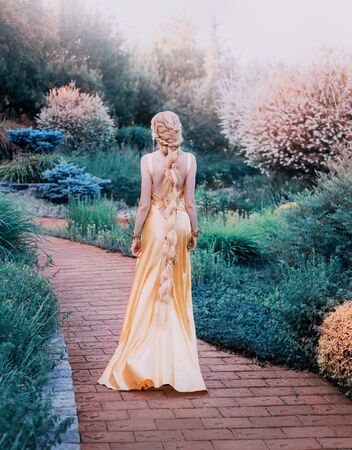 mysterious lady in chic yellow expensive luxury dress in magnificent garden, mysterious princess with long blond hair braided in hair stands with her back to camera, mystical fairy of nature, no faceの写真素材