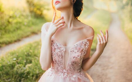 gentle elegant photo of girl with fair skin and dark hair, cute slender lady with bare shoulders twisted off straps of pink peach shiny dress, princess touches long neck with slender fingers, no faceの写真素材