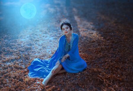 Young woman with the moon. Night autumn landscape. The Eastern Queen in a blue dress sits on the ground covered with fallen leaves in the moonlight. Tales of Shikhirizada 1001 nights. Art photographyの写真素材