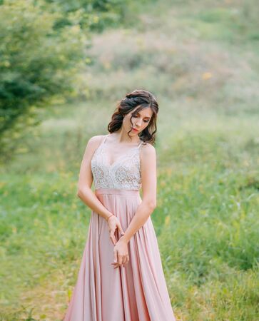 Cute attractive brunette woman enjoying nature in delicate elegant pink silk dress with white lace top. Image for party graduation prom ball stylish evening outfit celebration. Fashion glamor summer