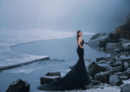 Gothic sexy woman vamp. tourist poses in creative long dress train on shore of frozen picturesque...