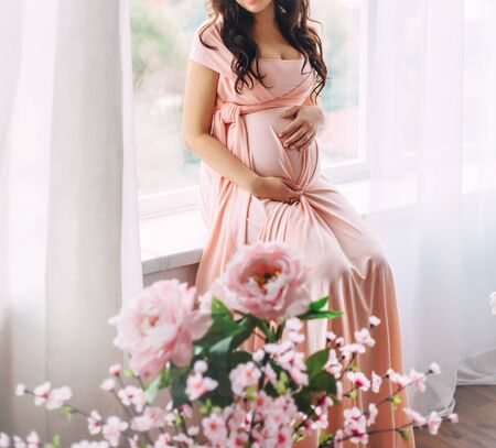 Happy pregnant brunette woman long hair peach dress sitting window in white room, love touches her belly. Bouquet pink flowers. concept natural cosmetics, expectation baby miracle. cropped smile faceの写真素材