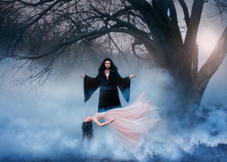 witcher men power magic spell raises woman air. Fluttering clothes mysterious cloak mantle hood. Backdrop autumn nature old tree white fog smoke cloud art creative levitation spirit long vintage gownの写真素材