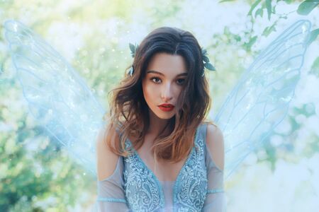 Fantasy closeup portrait attractive woman in image young fairy. carnival Costume blue dress transparent wings. face tender red makeup. Wavy Brunette hair Flutter fly in wind. Green nature backdropの写真素材