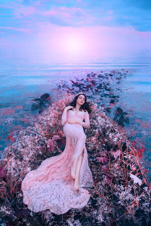 Fairy princess sleeping beauty in magical boat decorated with white red flowers tree branches. Trail of leaves water. long flowing hair brunette. pink long dress. Backdrop bright sky sunset sea wavesの写真素材