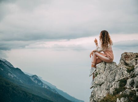Red hair girl turned away in pink suit sits on edge mountain rock on backdrop sky clouds mountains foggy haze. Woman enjoy beauty fairytale nature silence relaxation. Top Peak Monte Baldo Alps Italyの写真素材
