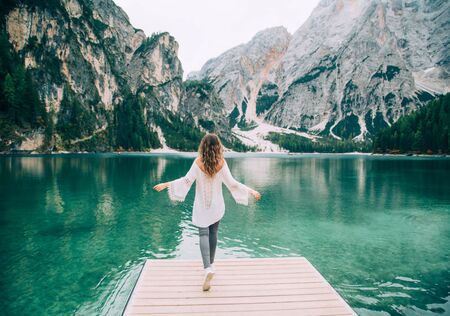 Romantic girl with long hair loose, turned away, walk spread hands on wooden pier Lake Braies. Tourist enjoy mysterious nature amazing Alpine mountains with green forest clear water silence relaxationの写真素材