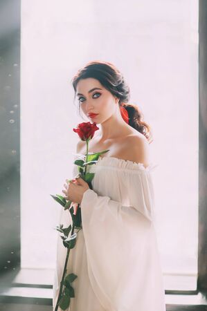 Portrait young beautiful bride woman. White medieval luxury sexy dress. bare shoulder. Brunette with long dark hair. Red rose in hands. backdrop sun light in window. Artistic wedding renaissance styleの写真素材