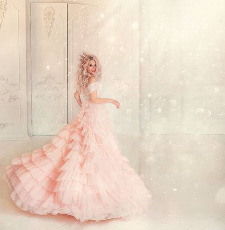 Happy young woman princess dancing in lush vintage pink dress. white royal room. Queen blond hair, golden crown. Light magic sparkles shiny. Carnival holiday invitation gesture. Background free spaceの写真素材