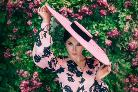 Artwork portrait young happy charming chic woman. Vintage stylish elegant retro dress. Brunette enjoy spring holiday on March 8, beautiful nature blooming pink flowers tree, rose bush. Hat wide brimの写真素材
