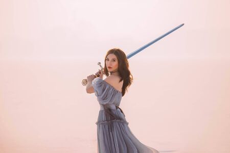 Art Portrait young beautiful brave warrior woman princess. Medieval queen holds steel silver magic sword. Concept Protection freedom. long hair retro vintage gray blue dress. White winter snow natureの写真素材