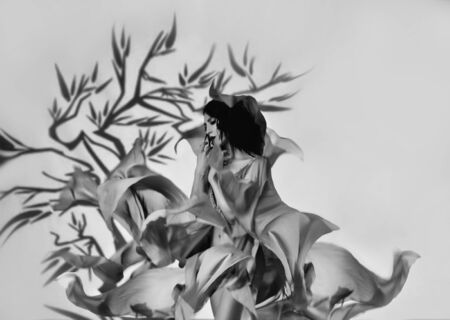Black white photo sexy beautiful woman in flower image. Airy dress silk fabric levitating flying in wind motion. Art studio shooting with projector spotlight. silhouette fantasy tree. magic fairy girlの写真素材