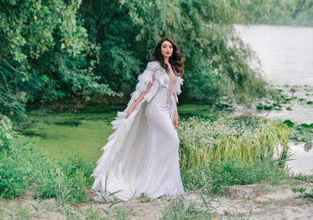 Beautiful Queen. charming woman bride walks on coast lake. Wedding outfit white long sexy dress, luxury cloak cape bird swan feathers. Brunette girl wavy loose hair. Backdrop summer nature green treesの写真素材