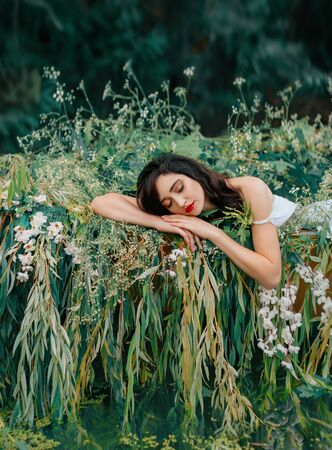 Young beauty sleeping woman resting in boat, decorated with willow branches, white flowers. Nymph girl closed eyes, enjoy silence summer nature river, water. Natural makeup red lips. Fantasy imageの写真素材