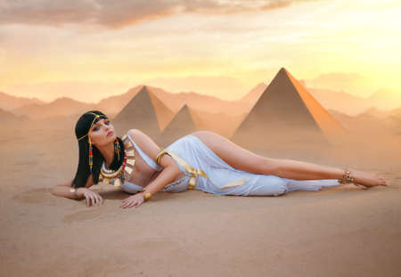 Egypt Style Rich Luxury Woman. Sexy beautiful girl goddess Queen Cleopatra lies on yellow sand desert pyramids. Art ancient pharaoh costume white dress gold accessories Black hair wig Egyptian makeupの写真素材
