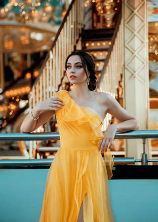 Young beautiful woman in a bright yellow evening dress posing against the backdrop of an amusement park and carouselの写真素材