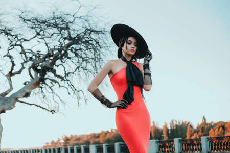 Elegant retro lady in a long red evening glamour dress. Young stylish trendy woman in a black hat...