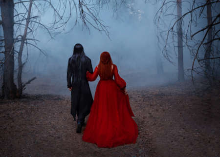 Blurred silhouette of a Gothic couple. Two people walks in the blue fog. A vampire man in a black tailcoat with long hair embraces a woman in a long red medieval dress. Deep forest, dark tree, smokeの写真素材