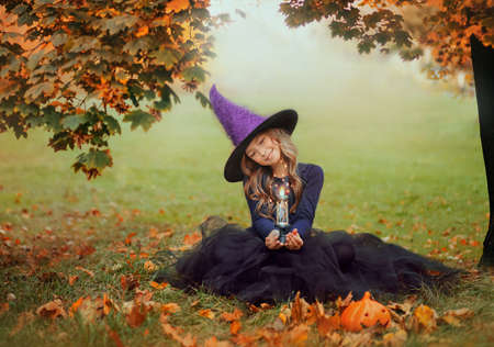A little beautiful girl in a witch costume sits on the green grass and holds a burning candle, magic...