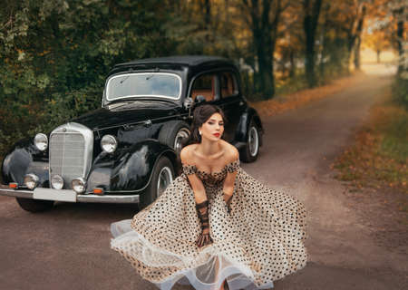 Young beautiful happy retro woman near the old car. Attractive elegant lady in vintage white polka black dot retro dress. Pin-up hairstyle. Cheerful girl standing on road. Autumn nature, yellow treesの写真素材