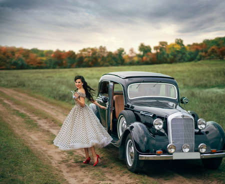 Young beautiful happy retro woman near the old car. Attractive elegant lady in vintage white polka black dot retro dress. Pin-up hairstyle. Cheerful girl standing on road. Autumn nature, yellow treesの写真素材