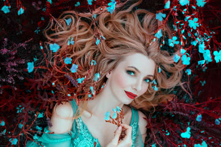 beautiful happy fantasy elf woman with blond hair lies dreams on grass, blooming meadow, blue flowers. Portrait smiling face, red lips elegant makeup. Romantic Girl princess resting enjoying lifeの写真素材