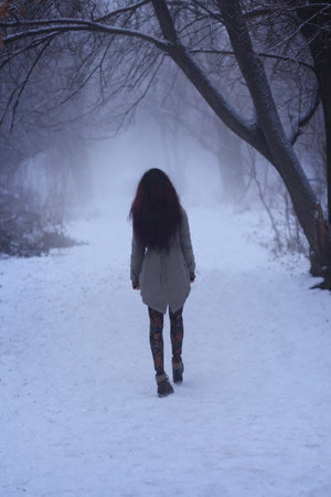 Silhouette of a sad young brunette woman walks away, along a path in evening misty park. Winter...
