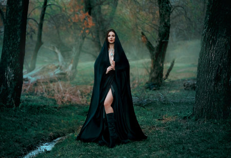 Mysterious fantasy gothic woman dark witch obsessed by evil. Girl demon vampire in black dress cape hood. walk in dark dense deep forest background, trees. Medieval queen in silk cloak, scarf posingの写真素材