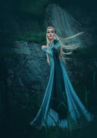 Beautiful fantasy woman elven goddess walks in nature mystical forest. Long creative vintage blue dress, sleeves. Blond hair fluttering in motion. Fairy cute face. Art Girl elf princess fashion modelの写真素材
