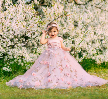 Young happy little girl fairy princess. Happy child face is smiling. Luxurious fluffy long dress for children, pink outfit. White flowers background trees spring nature. Fashion model, one year old.の写真素材
