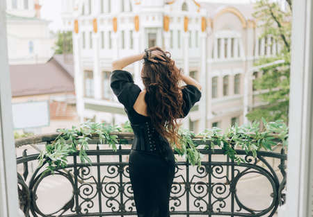 luxury woman in black evening dress stands on balcony looks at city. Back to camera rear view,...