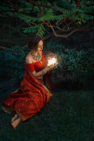 Young beautiful oriental indian fantasy woman sits on green grass, background night magic forest....
