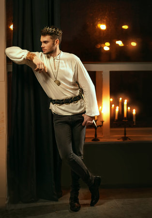 Portrait of handsome attractive man in image of medieval king. Vintage historic white shirt retro clothing gold crown on head. Dark gothic room, studio. Guy prince fashion model. Adult sexy guy knightの写真素材