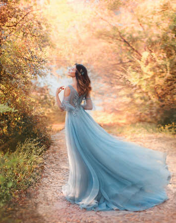 happy fairy woman princess in light summer blue dress standing in autumn park orange foliage tree. Lady walks fashion model. fantasy girl goddess. flying long hem of dress on wind. nymph of forestの写真素材