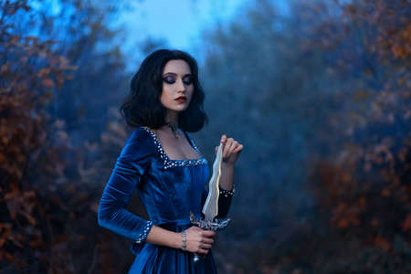 Fantasy medieval woman warrior queen holding dagger, knife in hands. Royal velvet vintage blue dress with pearls, girl princess vampire, brunette short hair. Nature forest dusk night, autumn seasonの写真素材