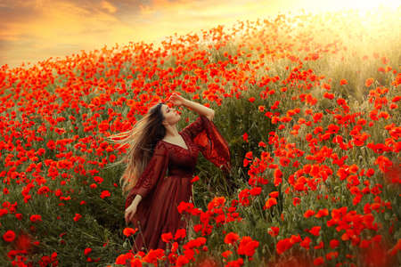 happy woman goddess walks in poppy field long hair flying in wind. Girl princess enjoys summer nature, red poppies flowers green grass, sun light shining, dramatic sky, sunset. vintage silk dressの写真素材