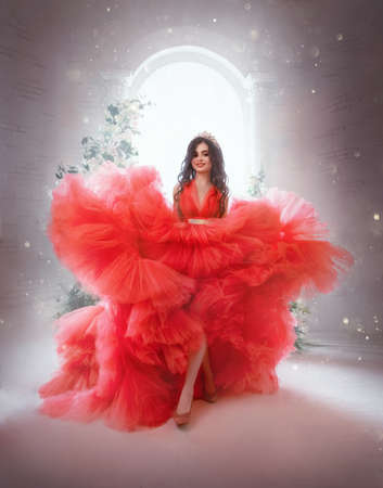 Happy cheerful fantasy princess in red ball gown. Queen woman smiling face. Carnival costume evening puffy lush long neon bright fuchsia color dress. Royal blue room, magical sun light from windowの写真素材