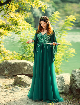 Fantasy warlike red-haired woman queen holding silver sword in hands praying before battle. Ritsa ritual of sacrifice. Green silk dress long elven sleeves, golden crown emeralds gems medieval styleの写真素材