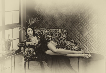 1920s old style sexy flapper woman lies on sofa, underwear and silk peignoir dress ostrich feather. girl holding glass of champagne in hand. luxury interior room. Black and white sepia vintage photoの写真素材