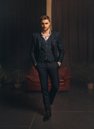 Portrait Handsome brutal man in classic strict three-piece suit jacket, trousers white shirt dark blue checkered vest. Successful businessman sexy guy stubble on face looks at camera. dark black roomの写真素材