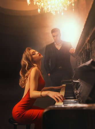 Luxurious sexy blonde woman pianist playing piano, party concert dark gothic scene, elegant man in black suit listens to melody music. Musician girl enjoys performance show. Red lips, dress bare backの写真素材