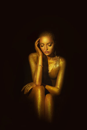 Portrait Closeup Beauty fantasy woman face in gold paint. Golden shiny skin. Glamorous fashion model girl image goddess in liquid gold. Professional metallic glowing luxury art makeup black backgroundの写真素材