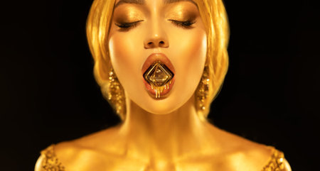 Portrait closeup Beauty fantasy woman sexy mouth hold diamond liquid gold drops drip on lips face in golden paint shiny skin. Fashion model sexy girl metallic makeup. creative jewellery. black studioの写真素材