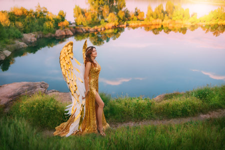 Art photo Fantasy woman goddess angel walks path blue sky reflection on lake water river, magic divine sun rays light. golden white wings long gold dress costume feather bird. Fairy girl sexy ladyの写真素材