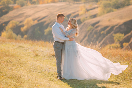 Couple in love together holiday wedding day, marriage concept. Happy man woman walking in park summer nature sun light green grass. Lady bride in long light gray dress, groom in business festive suitの写真素材