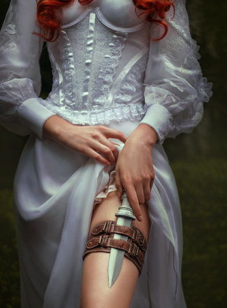 Fantasy red-haired dangerous woman medieval assassin queen with dagger in sheath on leg leather belt. White vintage old style dress. Girl princess warrior. Green summer nature trees. Hand hold knifeの写真素材