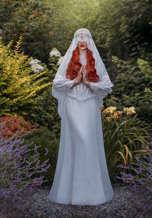 Fantasy woman in white vintage dress holy virgin prays face is hidden by silk veil. Red-haired girl nun queen. Medieval princess bride in garden green trees flowers. Mystical image sexy lady vampire.の写真素材